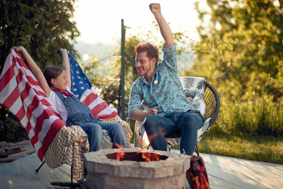 Cheerful little girl on an usa flag sitting next to a guy both on a deck with a bonfire