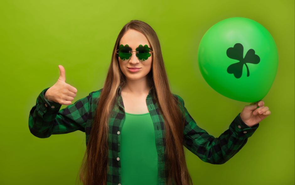 St. patrick's day young woman in green checkered plaid shirt with clover shaped glasses showing thumb up and holding air balloon with shamrock print isolated on green background
