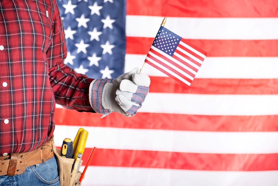 Worker with a tool bag and wearing a glove holding a small american flag with an american flag background
