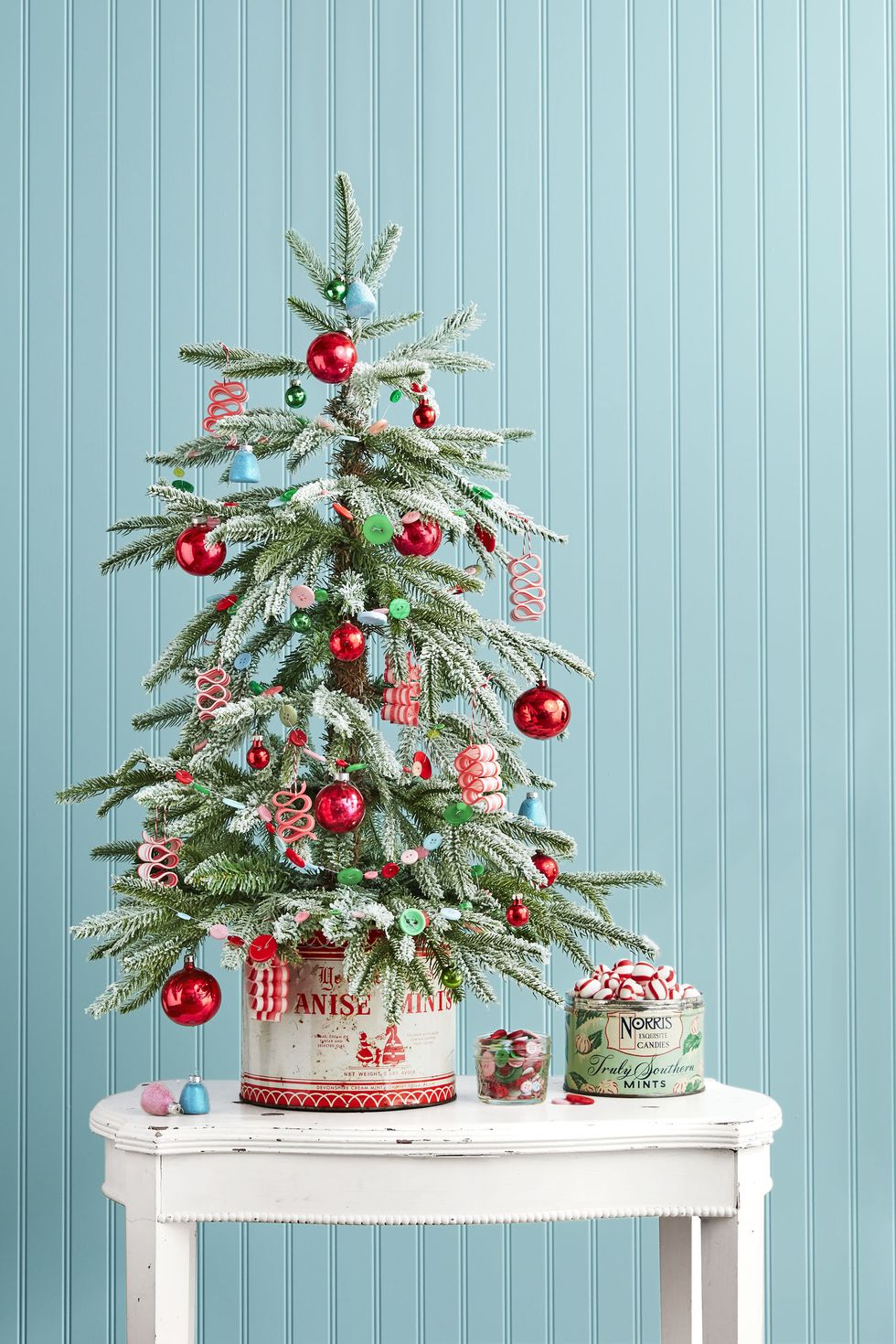 A small Christmas tree on a small table decorated with red and green ornaments against a light blue wall.