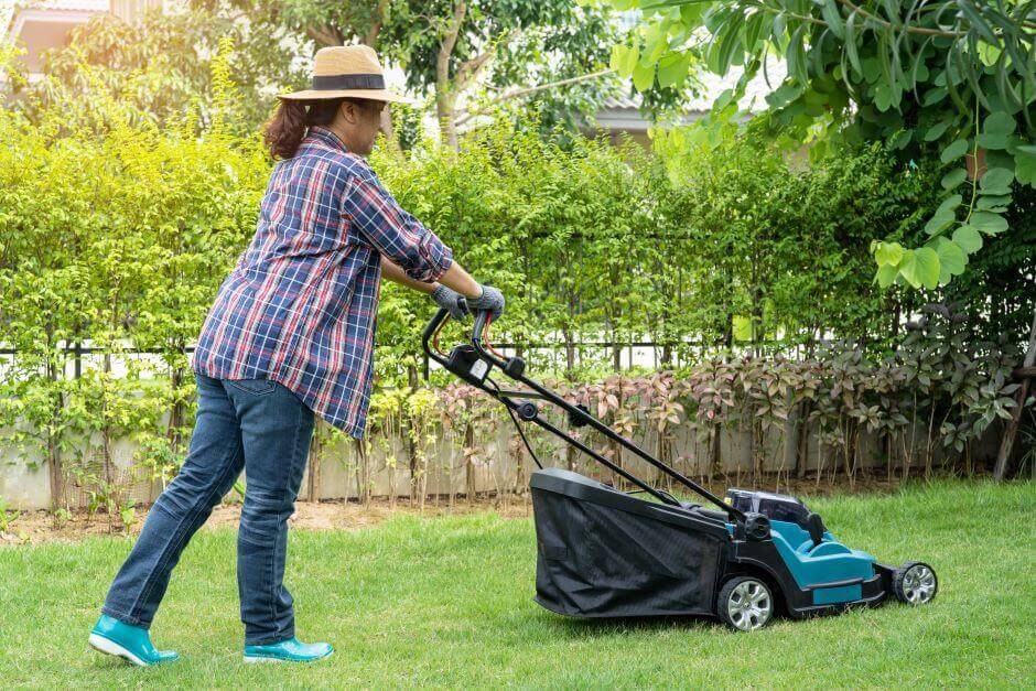 Woman using a hat and rain boots mowing the grass with a mower machine.