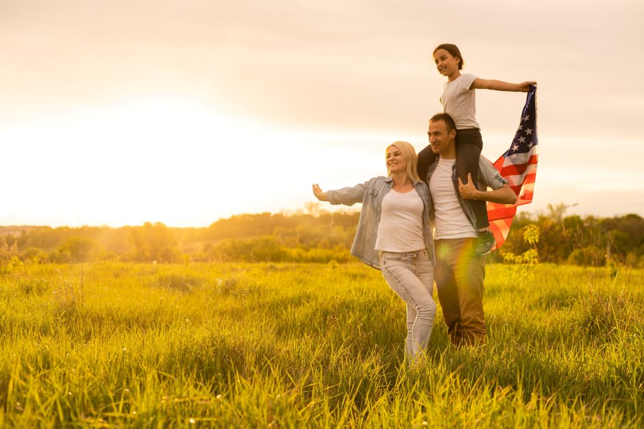 Happy family on a wheat field with the dad holding his daughter behind his back and the daughter holding an american flag in one hand