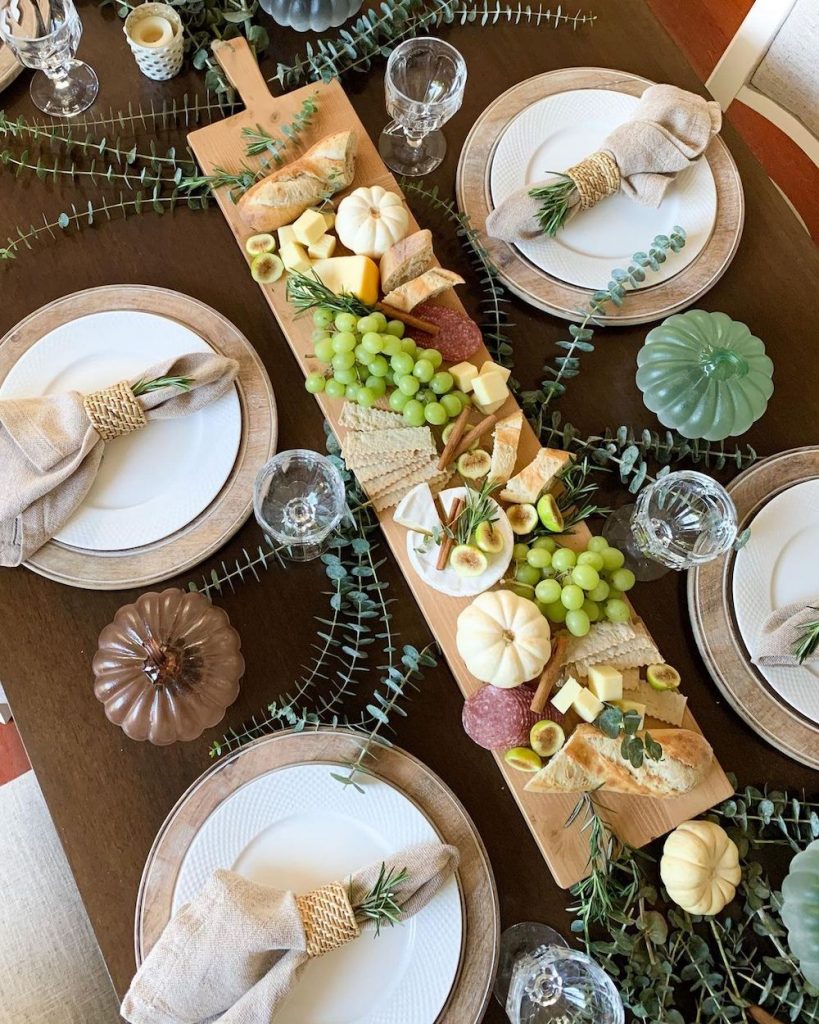A long cutting board placed in the middle of a dinner table topped with bread, cheese, green grapes, crackers, and decorated with small pumpkins.