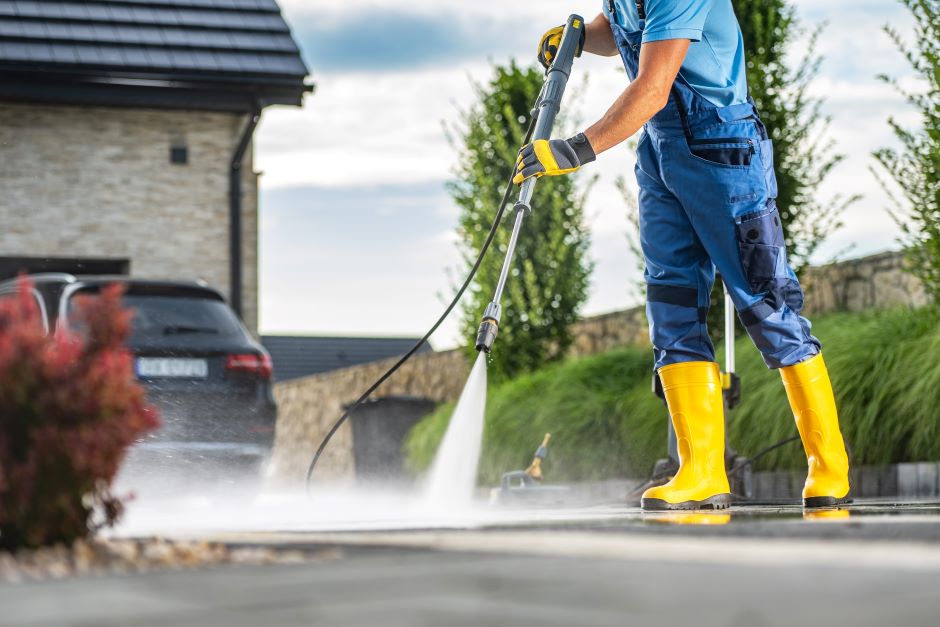 Man in overalls and protective boots, cleaning the sidewalk with a water jet. In the background, a house and a car