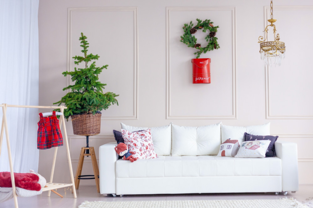 A simple living room area with a white couch decorated with Christmas-themed pillows, a small wreath on the wall, and a small pine tree on the side without ornaments.