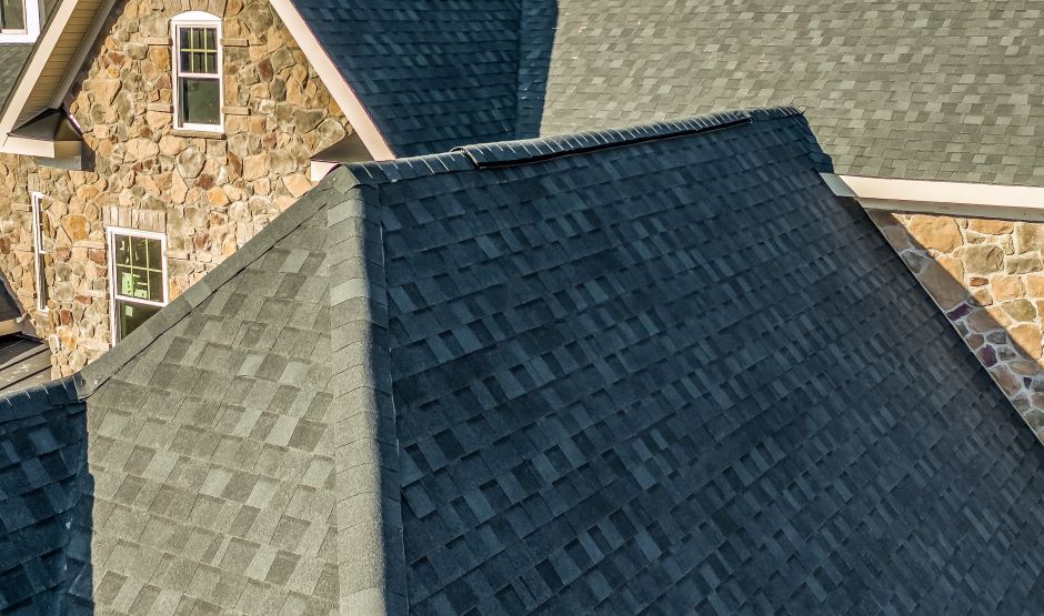 Roof of a house showing the ridge flashing system. In the background there is a house made of stones with two white windows.