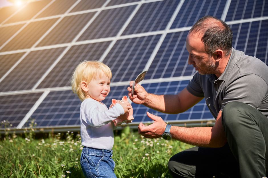 Adult male and child standing in front of solar panels, the child holds a piggy bank while the man deposits money