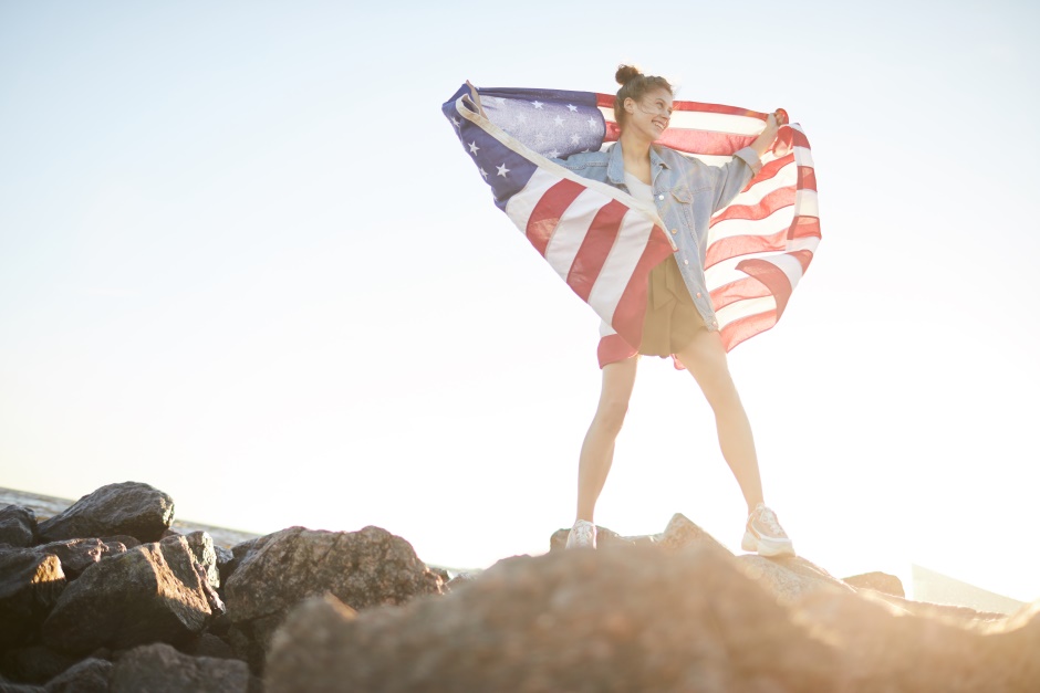 Happy excited young woman holding an american flag behind her back on a rocky beach terrain