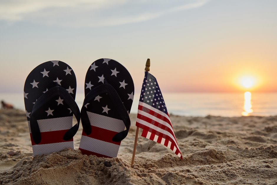 Flip flops painted with the american flag next to a small american flag buried in the sand on the beach with sea and sun in the background